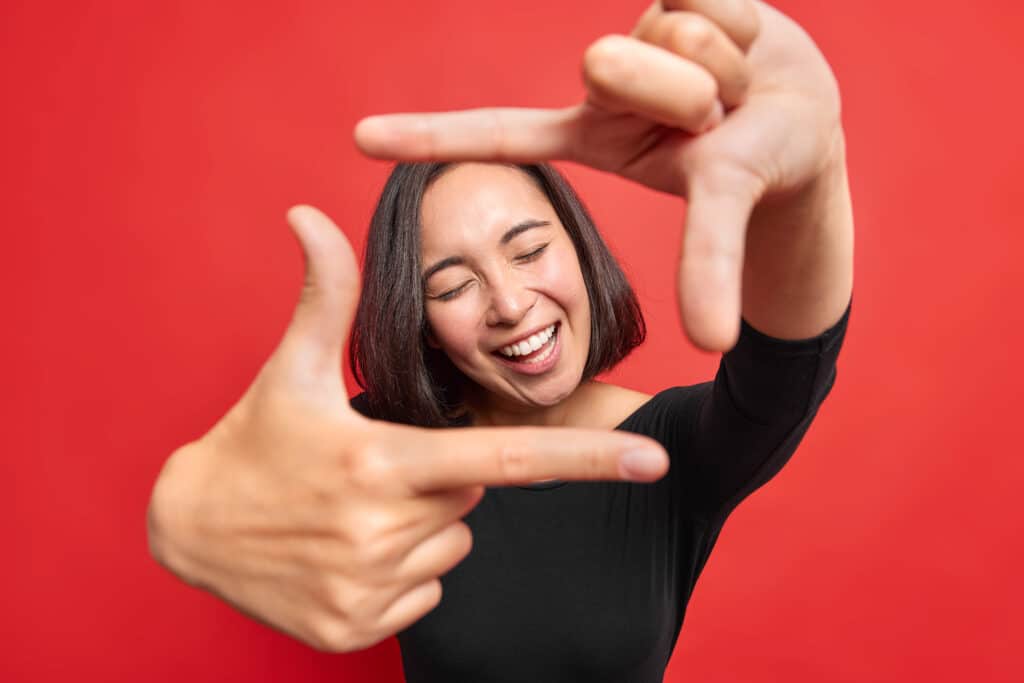 beautiful cheerful woman makes frame gesture smiles positively keeps eyes closed composes picture idea dressed in black jumper poses against vivid red background gazes at camera through hands